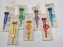 Six metallic tuning forks in blue, green, purple, gold, red, and turquoise, each placed on a beige fabric drawstring pouch with printed chakra symbols and names. The items are arranged on a white background with a faint 'COSMOCRAFT' watermark.